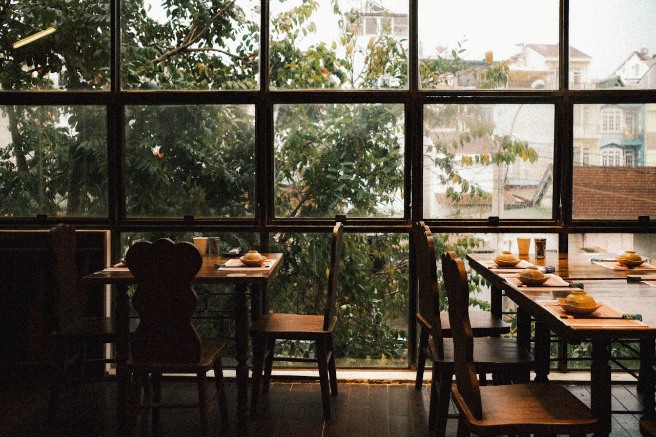 Warm and inviting restaurant interior featuring rustic wooden tables and chairs with a view of greenery through large windows.