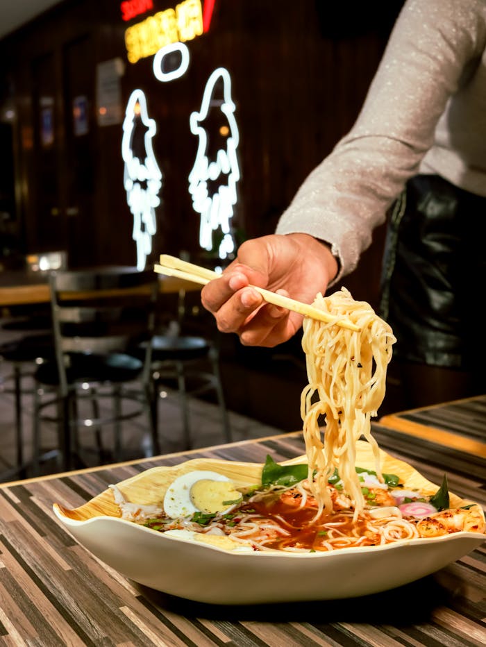 hero-img Close-up of flavorful ramen being lifted with chopsticks in a cozy restaurant with neon lights.